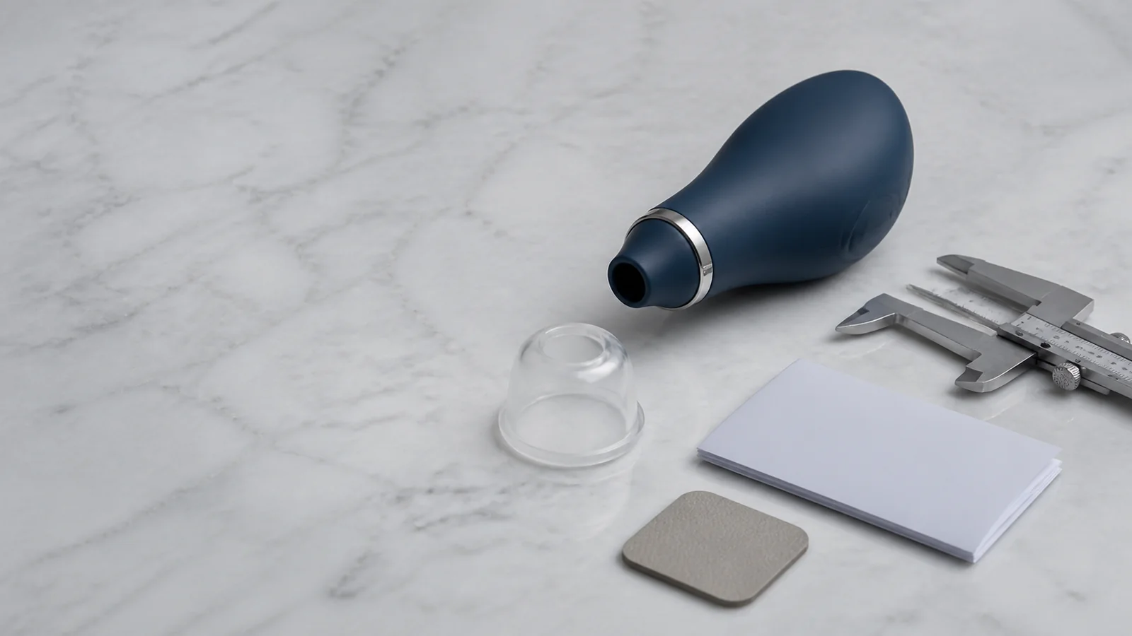 A compact midnight-blue suction-style wellness device beside a clear silicone cap and calipers in a clean studio.