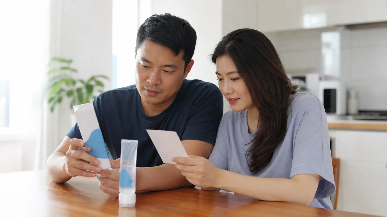 Two fully clothed Asian adults reviewing unbranded water-based lubricant packaging and a plain product information card at a kitchen table.